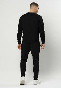 Bărbat stând cu fața spre spate, purtând hanorac negru și pantaloni, cu adidași albi, pe un fundal simplu și deschis la culoare.