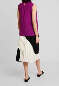 Lila ärmelloses Top mit einer Aussparung am Rücken, kombiniert mit einem plissierten Rock, der ein kräftiges schwarz-weißes abstraktes Muster aufweist. Schwarze Blockabsatzschuhe.