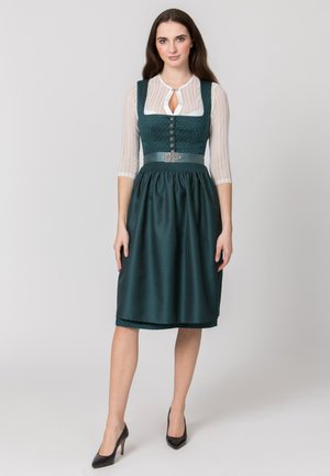 Frau trägt ein dunkelgrünes traditionelles Dirndlkleid mit weißer Spitzenbluse und schwarzen High Heels, steht vor einem schlichten Hintergrund.