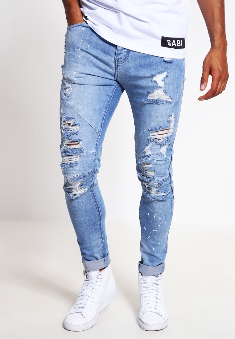 Jeans azzurri chiari con strappi grandi, aree sbiadite e macchie di vernice. Presentano una vestibilità slim e orli risvoltati, abbinati a sneakers bianche.