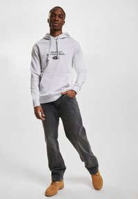 Grau Hoodie mit einer Vorder Tasche, schwarzer Logo Schriftzug, kombiniert mit dunklen Jeans und braunen Stiefeln. Das Material wirkt weich und hat eine lässige Passform.