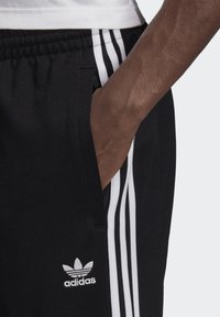 Čierne športové nohavice s bielym trojpásovým dizajnom, elastickým pásom a bočnou kapsou. Obsahujú logo adidas vyšívané bielou niťou.