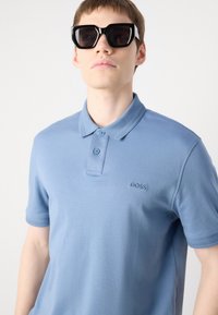 Polo de culoare albastru deschis, realizat din bumbac neted, cu un guler cu două butoane și logo brodat. Purtați cu ochelari de soare negri, îndrăzneți.
