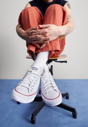 Weiße Leinensneaker mit roten Akzenten, niedrigem Profil, Metallösen und Gummisohlen, getragen mit weißen Socken und orangen Hosen, sitzend auf einem Stuhl.