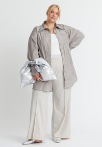 Helle graue, übergroße Shirtjacke mit spitzem Kragen, Knöpfen an der Vorderseite und einer Brusttasche. Kombiniert mit beigen, weit geschnittenen Hosen und metallischen Slippern.