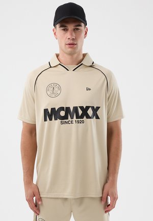 Mann har på seg en beige polotrøye med svart tekst "MCMXX SINCE 1920" og en rund emblem fra Buffalo, New York, beige shorts, og svart baseballcaps.
