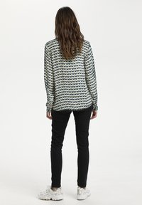 Camicia a maniche lunghe con un motivo geometrico blu, verde e crema; abbinata a pantaloni skinny neri e sneakers di colore chiaro.