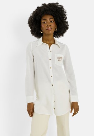 Femme aux cheveux afro portant une longue chemise blanche boutonnée avec broderie sur la poche et un pantalon crème, debout devant un fond blanc.