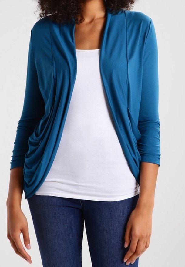 Femme portant un cardigan bleu à ouverture sur le devant, par-dessus un débardeur blanc et un jean bleu foncé, debout devant un fond uni.