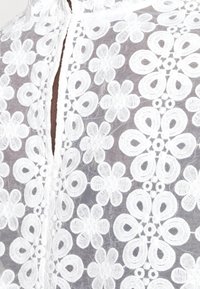 Tissu en dentelle florale blanche avec des motifs de fleurs à cinq pétales et bouclées brodés sur un fond gris transparent.