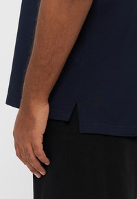 Marineblå kortærmet polo med struktureret stof, sideslidser ved sømmen og en afslappet pasform, kombineret med sorte bukser.