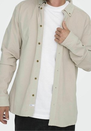 Homme portant une chemise beige clair boutonnée par-dessus un t-shirt blanc uni, ajustant le col de la chemise d'une main.