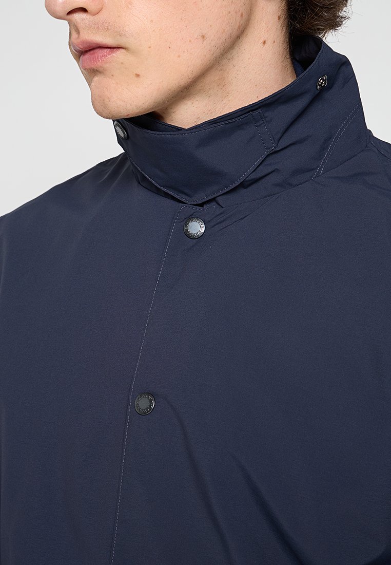 Dunkelblaue Jacke mit hohem Kragen, Druckknöpfen und einem strukturierten, wasserabweisenden Stoff. Nahaufnahme des Halsbereichs.