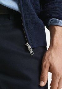 Suéter de punto azul marino con patrón de cuadros texturizados, presenta una cremallera de metal plateado, combinado con pantalones oscuros y una camisa azul clara debajo.