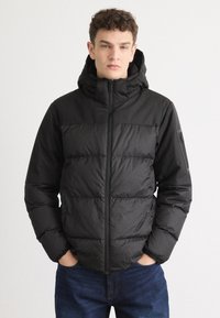Zwarte pufferjack met een rits aan de voorkant, capuchon en een gequilt ontwerp. Voorzien van elastische manchetten en een accentzak op de linkerarm.