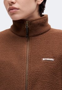 Κοντινό πλάνο ενός ατόμου που φορά καφέ fleece μπουφάν με φερμουάρ στον ψηλό γιακά και ορατό λογότυπο Columbia στο στήθος.