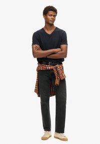 Camiseta negra de cuello en V, jeans grises y zapatillas beige. Camisa de cuadros atada a la cintura con tonos naranjas y rojos. Tejido suave y corte relajado.