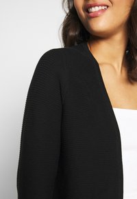 Svart stickad cardigan med en strukturerad design, öppen framtill och långa ärmar. Tyget verkar mjukt och kompletterar den vita toppen under.