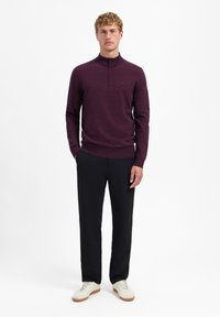 Merinowollpullover in Dunkellila mit Stehkragen und Reißverschluss, kombiniert mit schwarzen Hosen und weißen Sneakers. Glatte Textur, taillierte Passform.