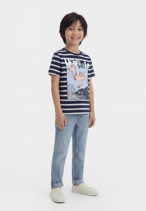Uśmiechnięte dziecko w granatowo-białej koszulce w paski z kreskówkowymi stworzeniami morskimi, jasnoniebieskich podwiniętych dżinsach i białych trampkach.