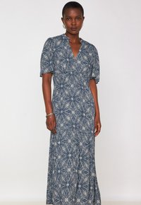 Femme portant une robe maxi bleue ornée de motifs floraux blancs complexes, avec des manches bouffantes courtes et un décolleté en V, se tenant devant un fond léger uni.