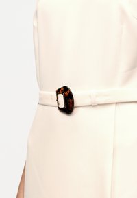 Λεπτομέρεια στη μέση ενδύματος κρεμ χρώματος με λεπτή ζώνη που διαθέτει μεγάλη ορθογώνια αγκράφα με μοτίβο χελώνας.