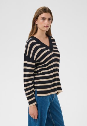 Femme debout, portant un pull rayé noir et beige à col en V et un jean bleu, regardant devant elle avec une expression neutre.