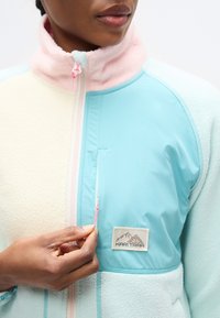 Ζακέτα με φερμουάρ από fleece και νάιλον σε παστέλ χρώματα: μπλε, ροζ και κρέμ. Διαθέτει λογότυπο και τσέπη με φερμουάρ με ροζ ταινία έλξης.