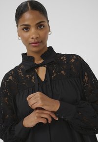 Blusa nera con maniche lunghe in pizzo floreale, colletto arricciato, scollatura a chiave con fiocco e dettagli a bottoni sui polsini.