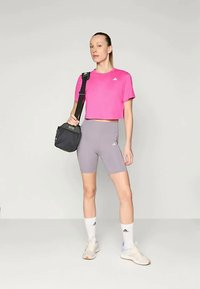 Naine seisab seljas roosa lühikese särgiga, hallid jalgrattapüksid, valged sokid, tennisetossud ning kannab musta õlakotti.