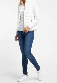 Person klädd i en vit långärmad skjorta, vit vadderad väst, blå tajta jeans och vita sneakers, stående med ett ben något böjt.