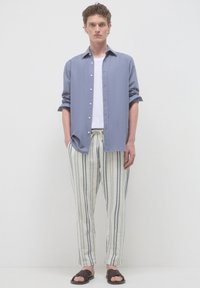 Junger Mann steht, trägt ein hellblaues, kurzärmliches Hemd über einem weißen Tanktop, cremefarbene gestreifte Hose und braune gewobene Sandalen.