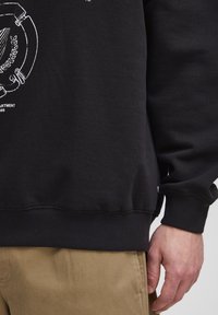 Sudadera negra con estampado gráfico en blanco que presenta un diseño circular y texto. Puños y dobladillo acanalados. Lleva encima un pantalón beige.