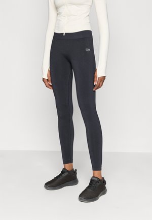 Sorte tætsiddende leggings lavet af strækbart materiale, med en høj talje og et lille logo på siden, parret med sorte sportssko.