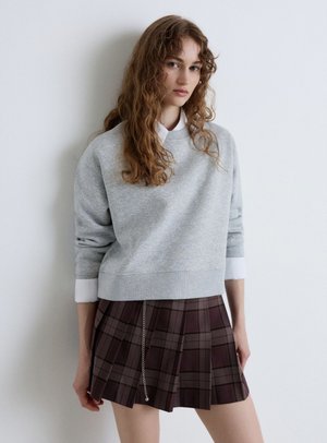 Jeune femme aux longs cheveux bouclés portant un sweat-shirt gris clair sur une chemise blanche et une jupe plissée marron à carreaux avec un détail chaîne, debout contre un mur blanc.