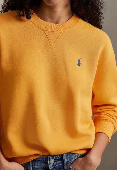 Żółta bluza z pulowerem z prążkowanymi mankietami i dekoltem, z małym niebieskim haftowanym logo na piersi oraz miękką fakturą.