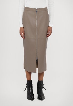 Mujer con falda midi color topo con cremallera frontal y abertura, combinada con botines negros y una blusa blanca de manga larga, de pie sobre fondo blanco.