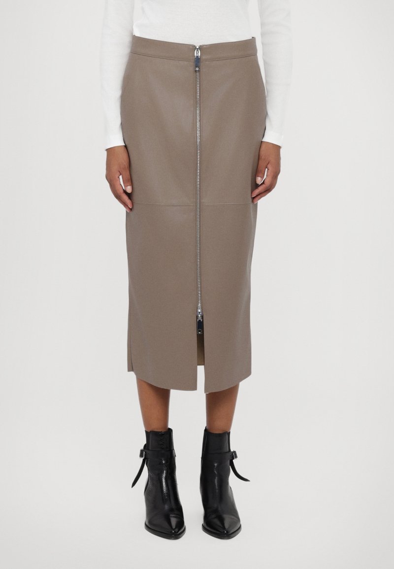 Femme portant une jupe midi taupe avec fermeture éclair sur le devant et fente, associée à des bottines noires et un haut blanc à manches longues, debout devant un fond blanc.