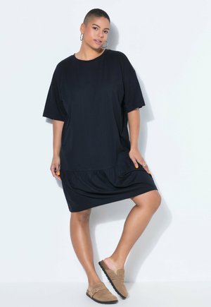 Kobieta o krótkich włosach, ubrana w luźną czarną sukienkę, z kolczykami w kształcie koła i beżowymi butami wsuwanymi, stojąca na tle gładkiej białej ściany.