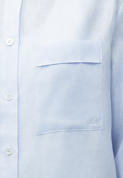 Φωτεινό γαλάζιο πουκάμισο με κουμπιά και τετράγωνη τσέπη στο στήθος με καπάκι και διακριτική κεντητή λεπτομέρεια "MM" κοντά στην κάτω δεξιά γωνία.