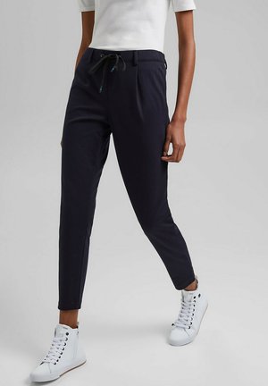 Kobieta w czarnych zwężanych spodniach z sznurkiem w pasie i białych wysokich trampkach, stojąca na jednolitym szarym tle.