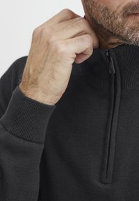Sudadera negra con cuello de media cremallera, puños acanalados y tejido texturizado. Una mano ajusta el cuello, destacando el detalle del cierre.