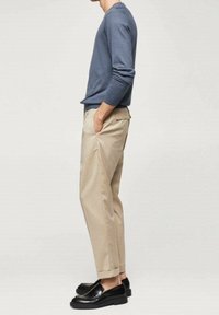 Ein Mann, der ein blaues Langarmhemd, beiges hochgekrempeltes Hosen und schwarze Slipper trägt, steht seitlich mit den Händen in den Taschen vor einem schlichten Hintergrund.