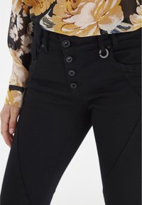 Jeans negros de cintura alta con cierre de cinco botones, trabillas para cinturón y una tela suave con detalles de bolsillos laterales.