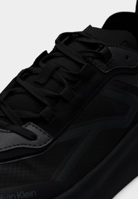 Κοντινό πλάνο μαύρου αθλητικού παπουτσιού Calvin Klein με υφή στην πλαϊνή επιφάνεια, κορδόνια στον ίδιο τόνο, ραφές και παχιά σόλα.