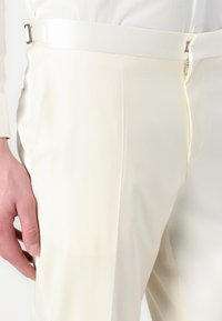 Světle krémové kalhoty s hladkým texture. Mají slim fit, boční zip a nastavitelné přezky na pase pro úpravu velikosti.