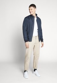 Navy lightweight bomber jacket with a stand collar, zip closure, and pockets, paired with beige trousers and white sneakers. Smooth texture.