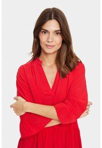 Robe rouge avec un décolleté en V, des manches trois-quarts et un ourlet évasé. Tissu doux offrant une texture lisse. Le modèle pose avec les bras croisés.