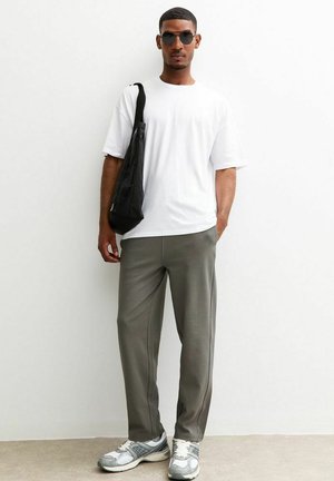 Camiseta blanca, pantalones de chándal verde oliva, bolsa de hombro negra y zapatillas grises. Modelo usando gafas de sol negras, de pie contra una pared lisa.
