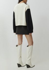 Pull en maille avec un corps crème et des manches noires, design court, poignets côtelés. Jupe mini noire associée à des bottes blanches jusqu'aux genoux avec des talons noirs.